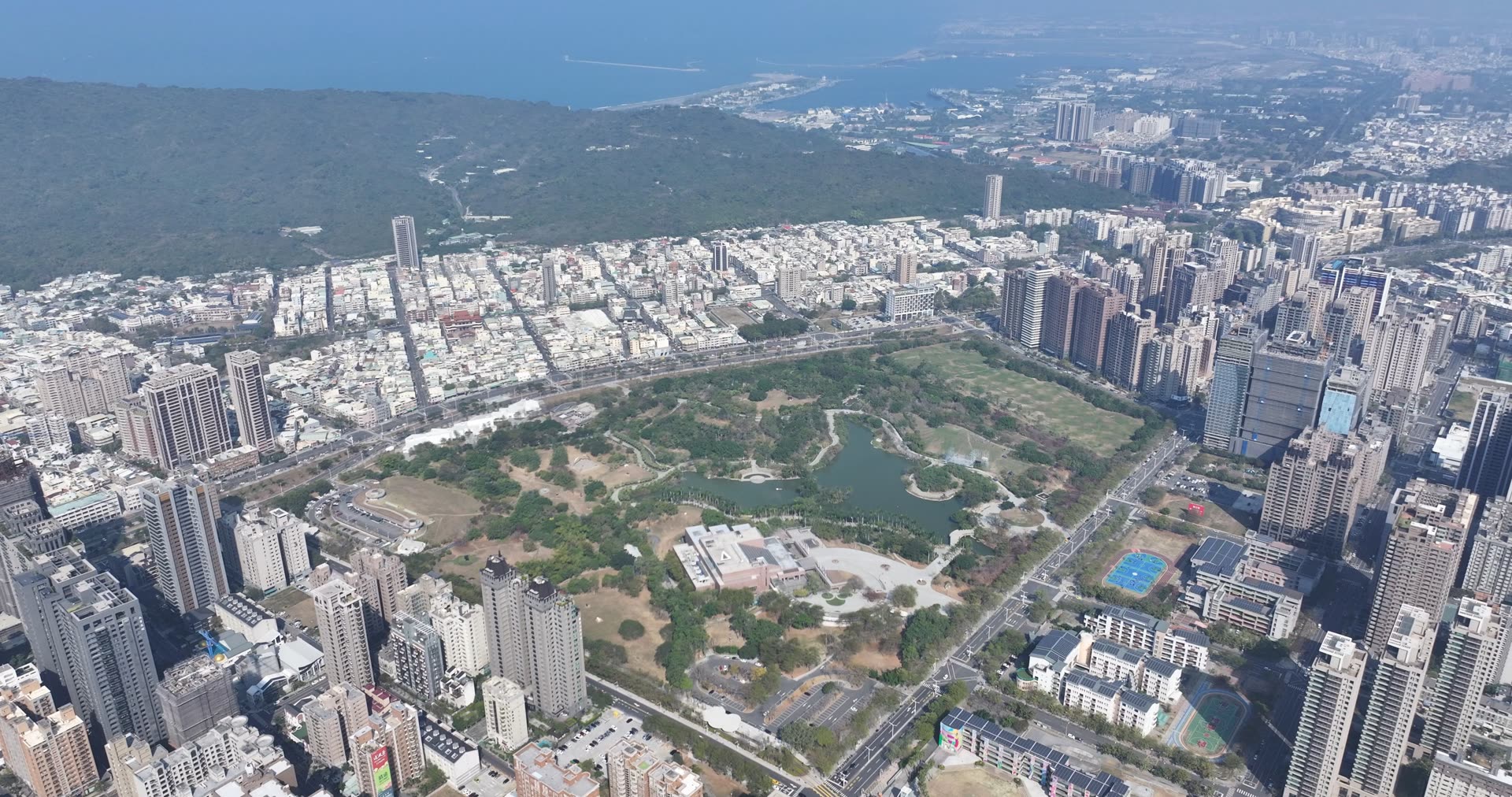 空拍鏡頭俯瞰高雄美術館特區的大片綠地、湖泊與城市天際線,遠方可見港灣,展現高雄宜居生活樣貌